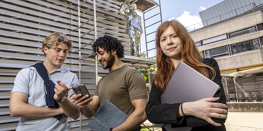 Zwei Studenten unterhalten sich am Campusgelände, daneben steht selbstbewusst eine Studentin.