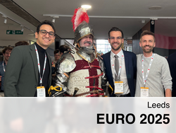Luca Santosuosso, Felix Auer und Robert Gaugl pose with a person in full medieval armor, complete with a helmet and plumed crest, at the EURO 2025 conference in Leeds. All three men are wearing conference badges.