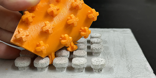 Ein quadratisches, oranges Plastickstück mit Noppen, die sich in ein entsprechendes Gegenstück auf einem Betonteil verhaken.
