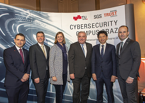 Five men and a woman are standing in front of an expo wall, on which the logos of TU Graz, SGS and the URL of the Cybersecurity Campus Graz can be read.