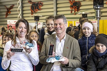 A woman and a man hold small hydrogen cars in their hands. Children stand around them.