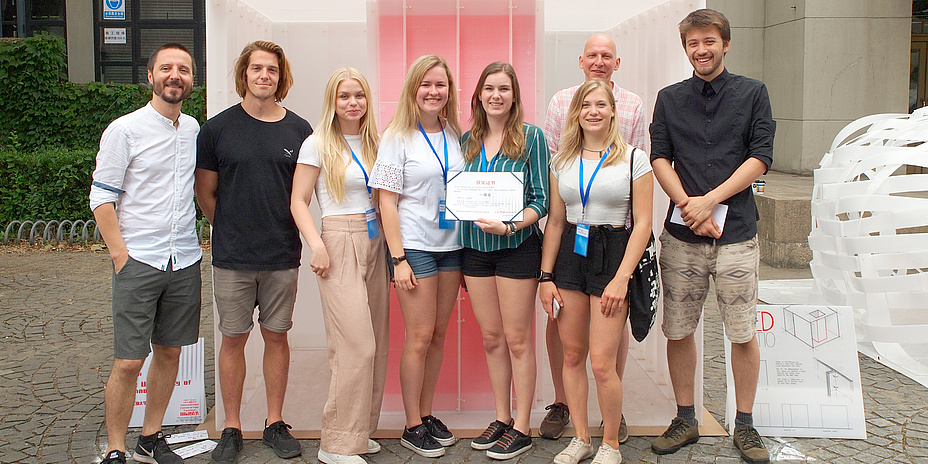 Two male students, four female students and two supervisors, all dressed in summer clothes, in front of a small pavilion they built themselves, next to which on the ground are the design drawings on a small poster