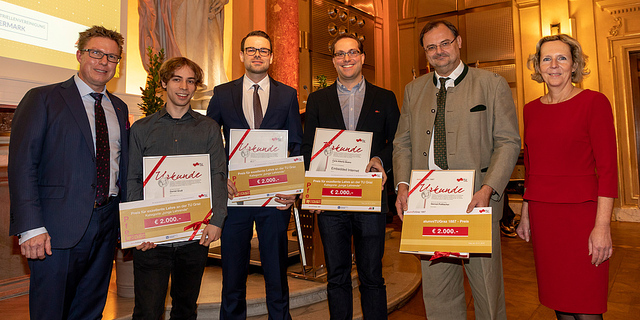 Six persons standing next to each other and those four in the middle holding certificates in front of their bodies into the camera can be seen.
