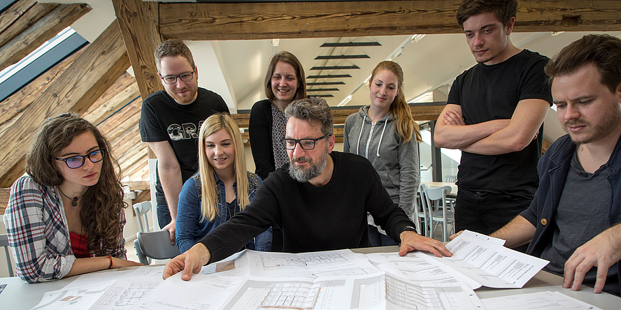 Universitätsassistent Armin Stocker sitzend an einem Tisch mit Entwurfszeichnungen umringt von sieben Studierenden vor der renovierten Dachbalkenkonstruktion in den TU Graz Masterstudios.