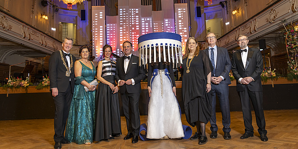 Sieben festlich gekleidete Menschen in einem Festsaal, in der Mitte eine kostümierte Person.