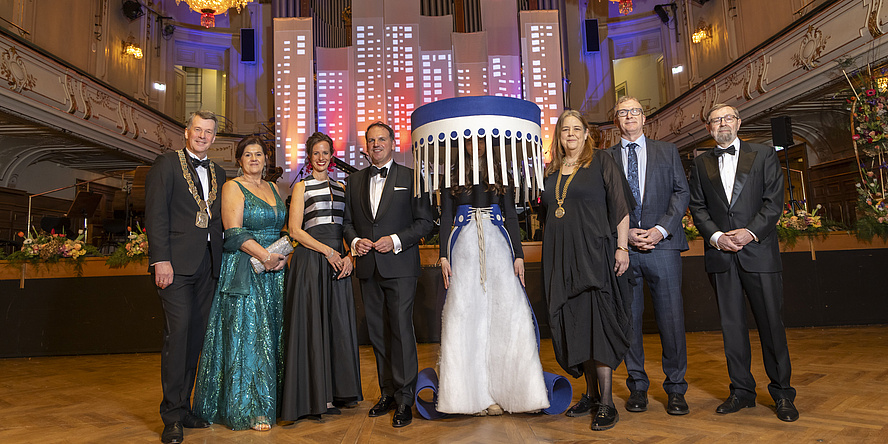 Sieben festlich gekleidete Menschen in einem Festsaal, in der Mitte eine kostümierte Person.