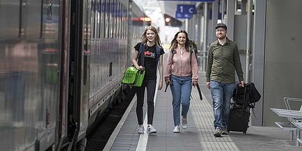 Two young women and a young man with luggage walk next to a train on a railway platform