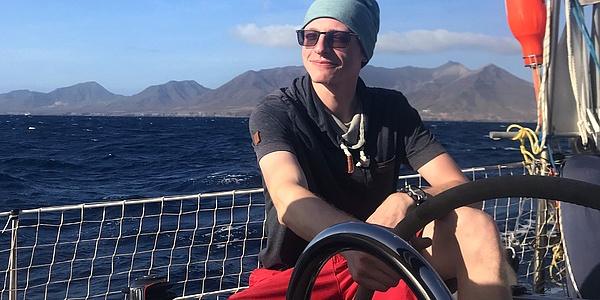 Man sitting on sailboat with sea and mountains in background.