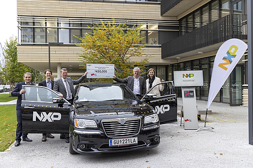 Five people, four men and one woman, stand next to a car and hold up a cheque with a value of 450,000 US dollars