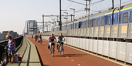 Bicyclists dressed for summer on the city bicycle path parallel to a moving suburban train on a rail bridge.