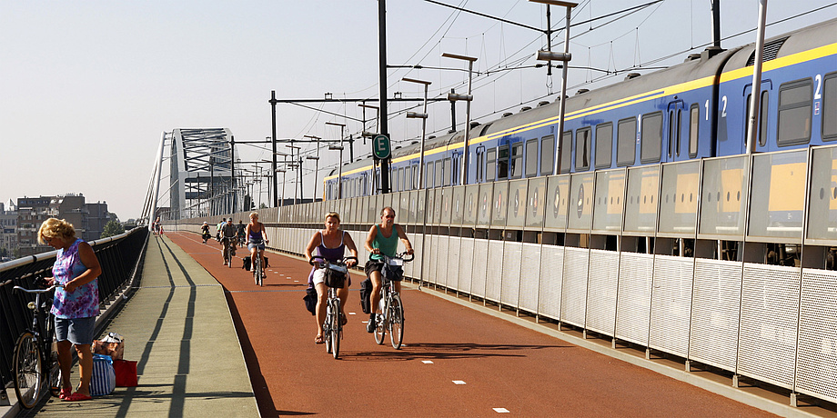 Bicyclists dressed for summer on the city bicycle path parallel to a moving suburban train on a rail bridge.