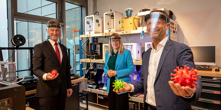 Eine Frau im türkisen Blazer und zwei Herren im Aanzug vor einer Galerie von 3D Druckern im Labor