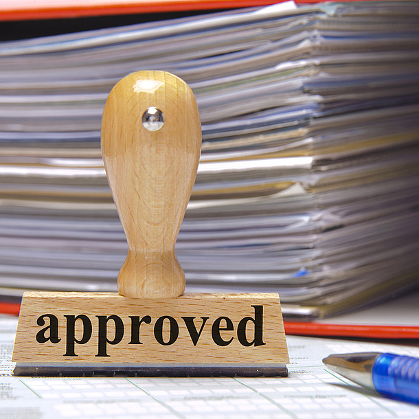 Ein Stempel mit der Aufschrift "approved", dahinter ein Stapel Papier. Bildquelle: Wolfilser – Fotolia.com