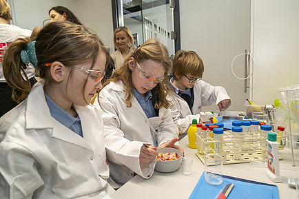 Drei Kinder in Labormänteln und mit Schutzbrillen.