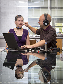 A man with headphones is working on a small device that a woman is wearing around her neck.