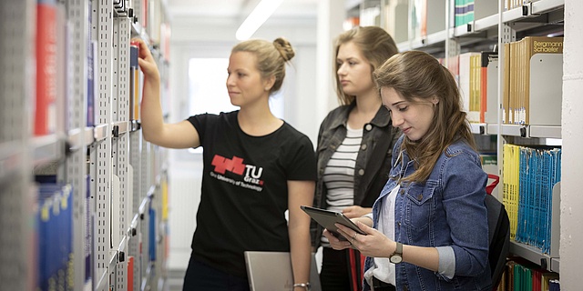 Drei Studierende stehen vor einem Bücherregal.