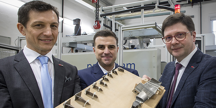 Christof Sommitsch, Sergio Amancio and Franz Haas. They are holding a brick of wood and several metalic objects. Christof Sommitsch is wearing short brown hair, a black suit, blue tie and white shirt. Sergio Amancio wears short, brown hair, a blue suit and white shirt. Franz Haas wears brown hair, red glasses, blue suit, white shirt and red tie.