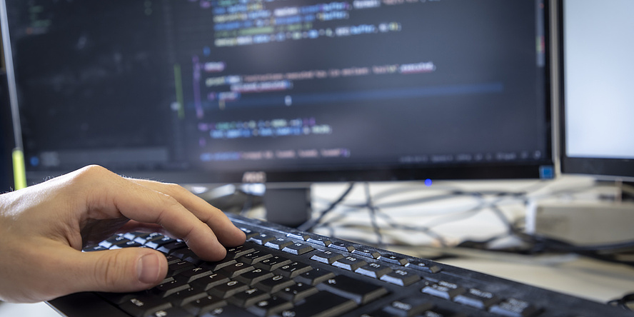 A hand types on a computer keyboard, a monitor with computer code can be seen in the background.