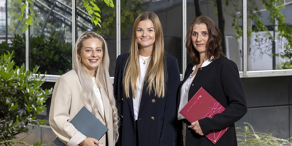 A blonde woman in a white jacket, a blonde woman in a dark jacket and a dark-haired woman in a blue jacket stand outside and look into the camera.