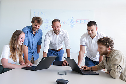 Five people are at a white table. On the far left, a woman is sitting at a laptop; on the far right, a man is sitting at a laptop. Three men are standing between them. The man in the middle is looking at the camera; the two people on the left are looking at the laptop on the left; the two people on the right are looking at the laptop on the right.