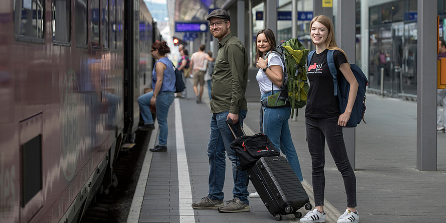 Two female and a male student carrying a trolley case and backpacks ready for departure on a platformat at the train station in Graz.