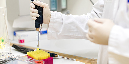 A laboratory situation, a hand with a research pipette docking to a small yellow measuring cylinder. At the table, further laboratory material can be seen, the person wears a white coat.