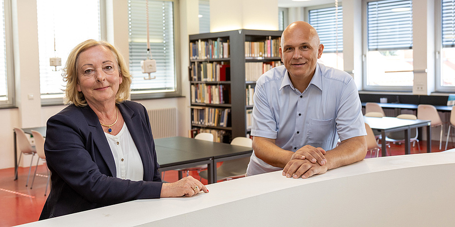 Eine Frau und ein Mann stehen in einer Bibliothek, im Hintergrund ein Bücherregal.