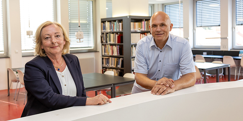 Eine Frau und ein Mann stehen in einer Bibliothek, im Hintergrund ein Bücherregal.