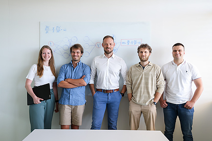 Five people are leaning against a white board. On the far left is a woman with a laptop under her arm, and to her right are four men. They are all smiling at the camera.
