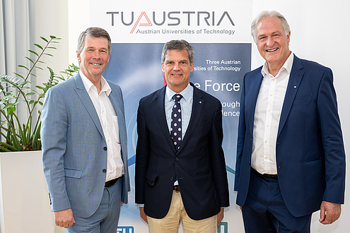 Three men are standing in front of a banner and are looking into the camera.