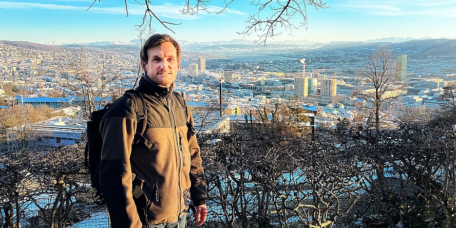 Ein Mann mit dunklen Haaren blickt in die Kamera. Er steht im Freien und es scheint kalt zu sein. Im Hintergrund sieht man eine Stadt. 