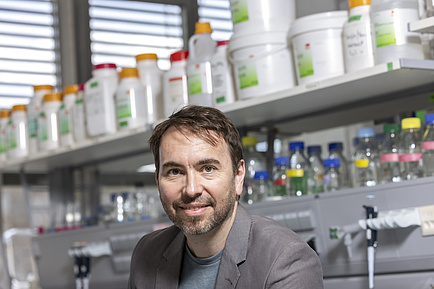 A man smiles at the camera. Behind him, white plastic containers with labels can be seen. It looks like a laboratory.