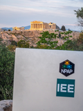 Ein Laptop mit den Aufklebern „NPAP“ und „IEE“ steht im Vordergrund, dahinter die Akropolis mit dem Parthenon in Athen, umgeben von Bäumen und Felsen.