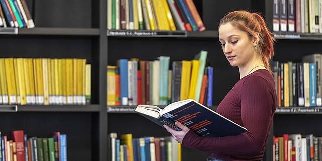 One person stands in front of a bookshelf and holds a book.