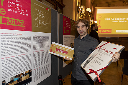 A man is holding a tribute in his right and left hand. To the left of him is a poster wall showing a poster in A1 format.