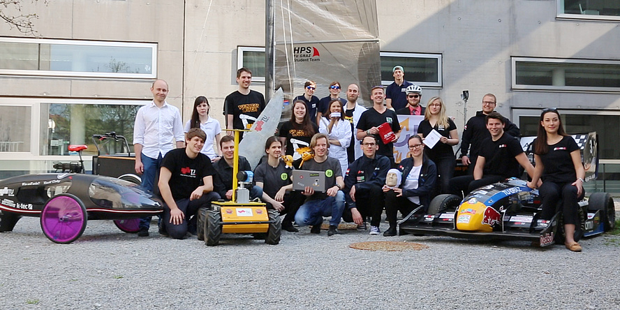 Gemeinschaftsbild von Studierenden aus Teams der TU Graz mit Rennautos, Roboter, Segeljolle und mehr.