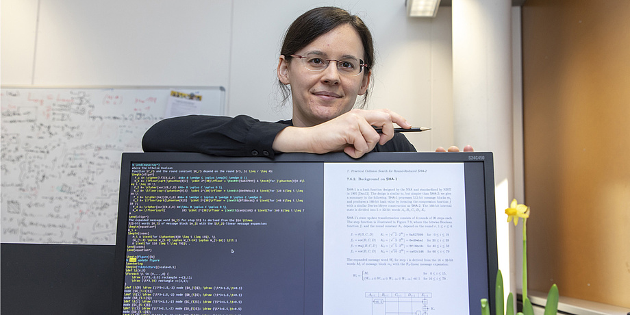 A woman leans on a computer monitor, holds a pencil in her hand and smiles into the camera