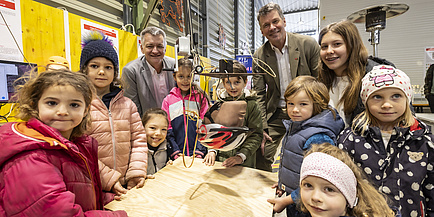 Zahlreiche Kinder und zwei Männer stehen um Holzplatte, über der ein künstlicher Kopf mit Fahrradhelm hängt.