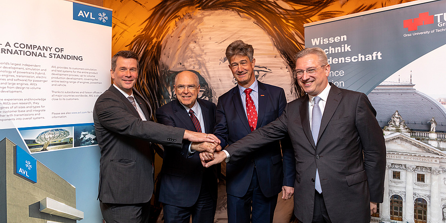 Four men in suits stand in front of the two roll-ups of AVL and TU Graz and cross their hands. 