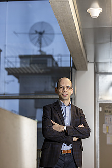 A man stands in a corridor with his arms crossed; a large antenna is visible through a window in the background.