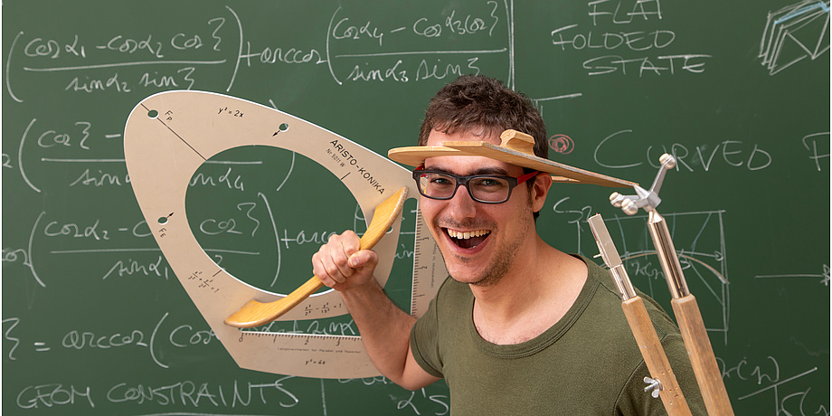 Ein lachender Mann im grünen T-Shirt steht vor einer grünen Tafel, auf der mathemaitsche Formeln geschrieben stehen und hält einen überdimensionalen Zirkel und eine Schablone für Kegelschnitte in den Händen.