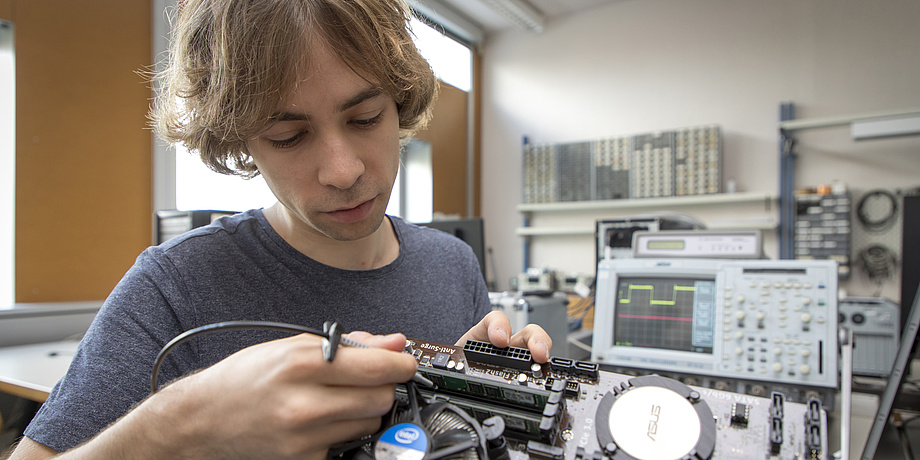 Ein blonder junger Mann sitzt an einem Tisch und arbeitet am Innenleben eines Computers.