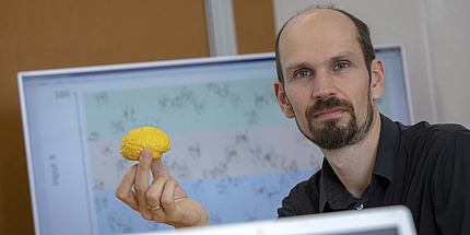 A man looks into the camera. He is holding a small, yellow model of a brain.