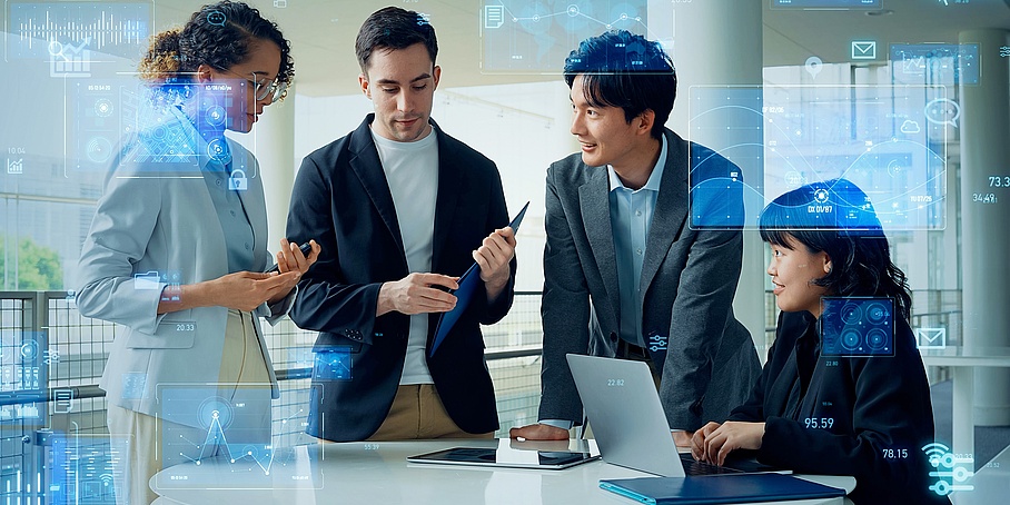zwei junge Frauen – eine steht und eine sitzt vor einem Notebook – und zwei junge Männer –stehend – arbeiten im Team, Business-Kleidung, Überblendung teilweise mit transparenten digitalen Bildschirmen