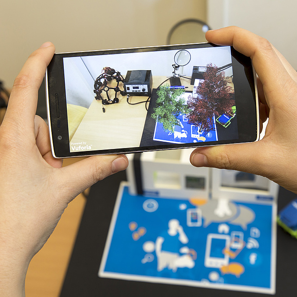 Two hands hold a mobile phone, on the screen you can see models standing on a desk.