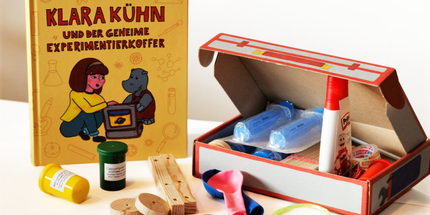 Gelbes Buch mit der Aufschrift „Klara Kühn und der geheime Experimentierkoffer". Daneben eine offene Kartonbox mit Klebstoff, Luftballonen, kleinen Dosen und Holzstäbchen.