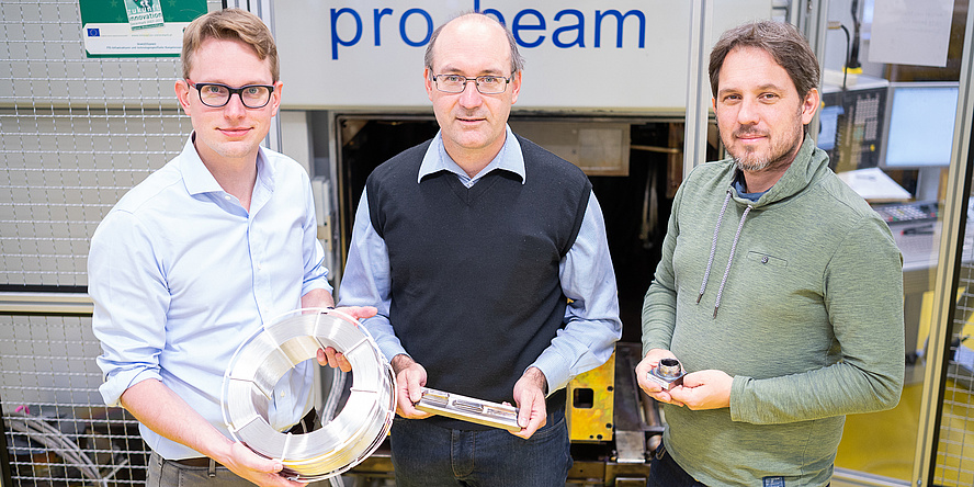 Three man in a lab. Florian Pixner wears blond hair, glasses and a blue shirt. He is holding a spindle of white cable. Norbert Enzinger wears glasses and a blue shirt. Fernando Warchonicka wears short brown hair, a beard and a green jumper.  