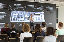 Lecturer in front of a large video wall, the seated audience can be seen from behind.