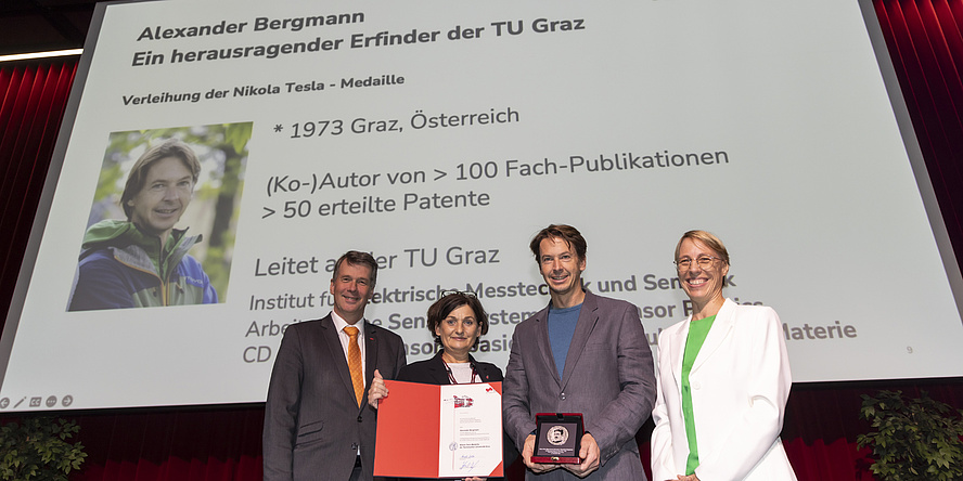 Zwei Männer und zwei Frauen stehen vor einer Leinwand und lächeln in die Kamera. Ein Mann hält eine Medaille