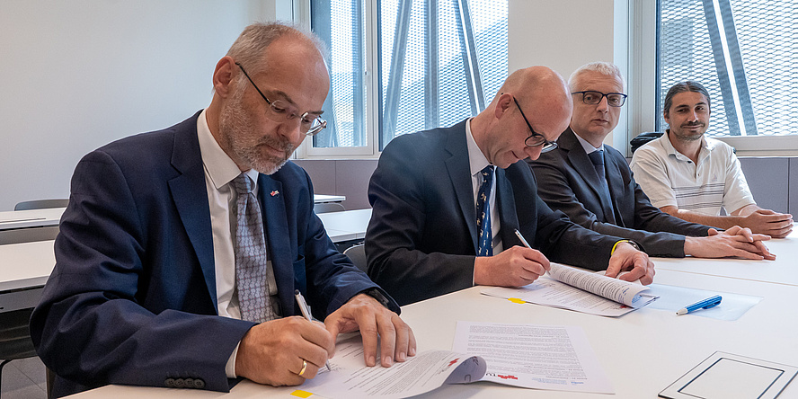 Vier Männer sitzen an einem Tisch. Die beiden auf der linken Seite unterzeichnen gerade ein Schriftstück, die zwei daneben sehen dabei zu.